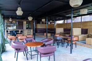 A café with round tables and cushioned chairs, a bar area with shelves, and a covered seating area with wooden partitions. - The Country Inn by Smart Haven (Kigali)