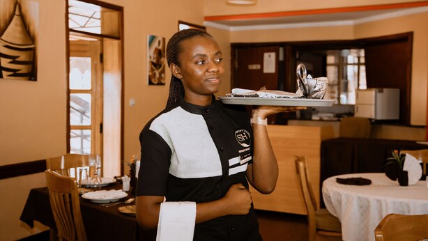 A dining area with tables set for meals, a waiter holding a tray with dishes, and a kitchen area in the background. - The Country Inn by Smart Haven (Kigali)