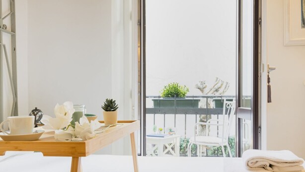 A balcony with a view of a garden, a table with a cup and flowers, and a stack of towels.