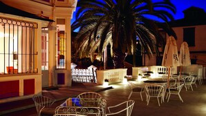 Un espacio al aire libre con sillas y mesas de metal blanco, una palmera, y un edificio con ventanas iluminadas y un balcón.