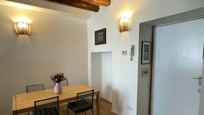 Uma sala de jantar com mesa e cadeiras de madeira, duas luminárias fixadas na parede, uma porta branca e um quadro emoldurado.