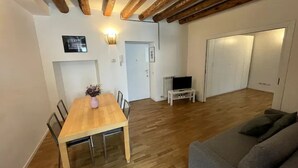 Uma área de refeições com uma mesa e cadeiras de madeira, um sofá, uma televisão sobre um móvel e uma porta que dá para outro espaço.