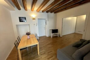 A dining area with a wooden table and chairs, a sofa, a television on a stand, and a door leading to another room.