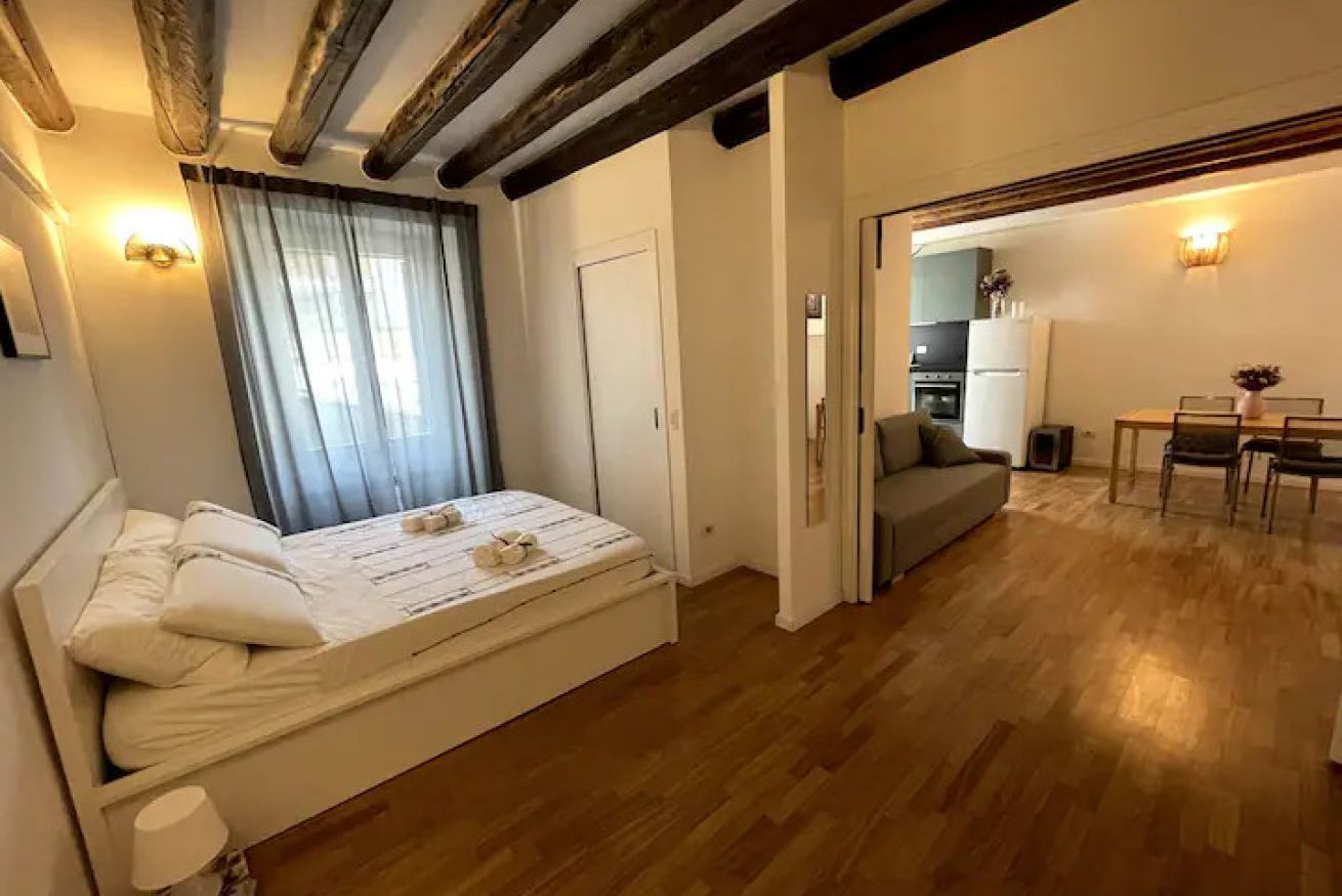Un dormitorio con cama, mesita de noche, ventana con cortinas, puerta y vistas a un comedor con mesa y sillas.