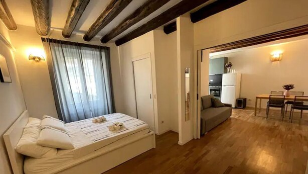 Um quarto com cama, mesa de cabeceira, janela com cortinas, porta e vista para uma sala de jantar com mesa e cadeiras.