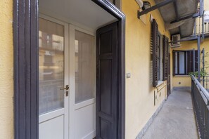 Un pasillo estrecho con una puerta cerrada, una ventana con persianas y un pequeño balcón con barandilla.