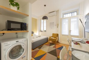 Habitación de hotel compacta con lavadora, microondas, cama, silla, mesa de comedor y ventana con persianas.