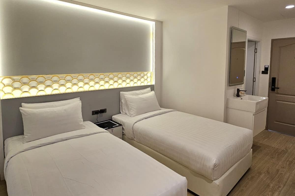 Une chambre d’hôtel moderne avec deux lits simples, une table de chevet, une commode, un bureau intégré, un miroir et une salle de bain avec un lavabo et un miroir.