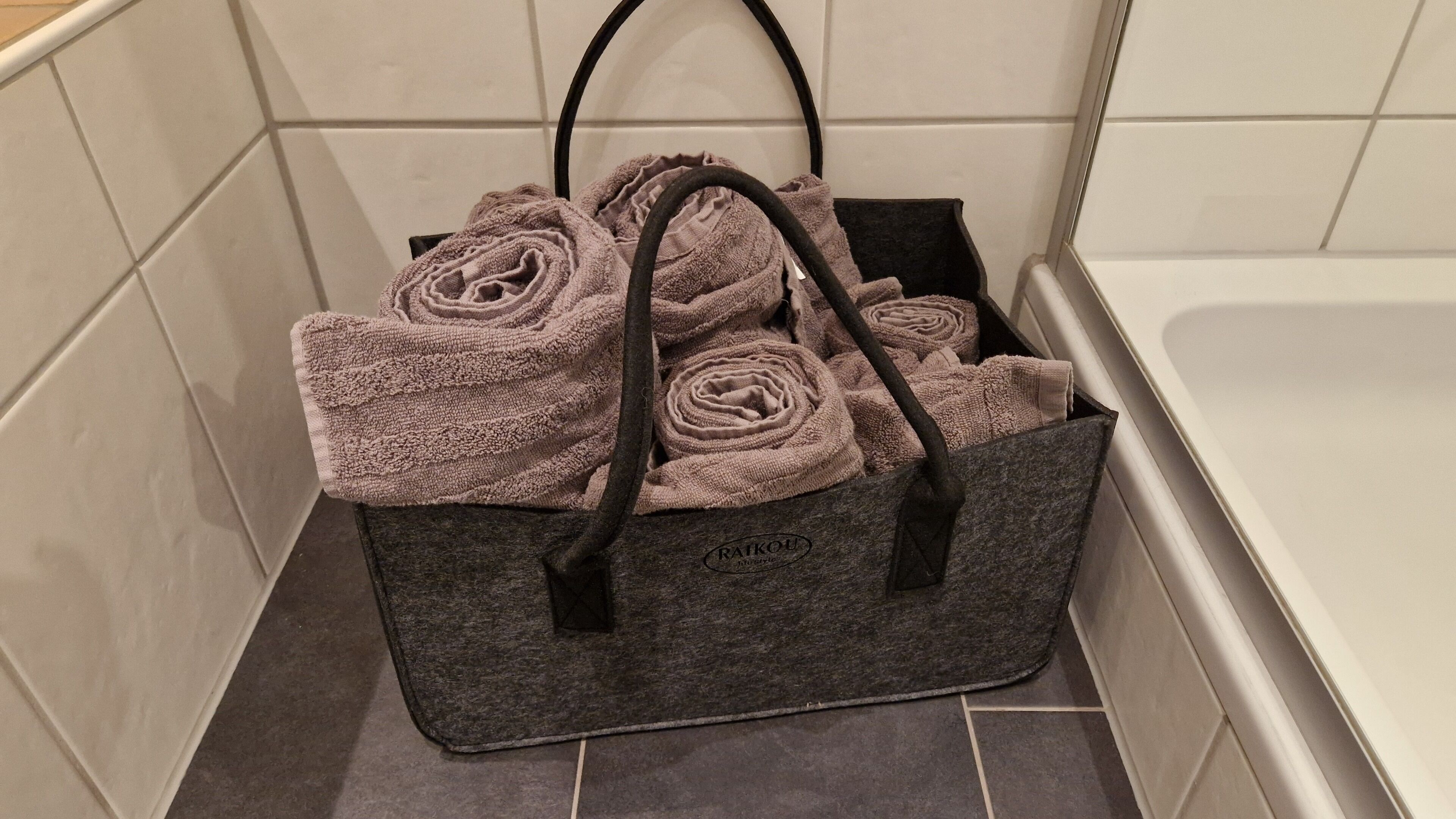 A dark gray tote bag with handles, containing several rolled towels, placed on a tiled floor in a corner.