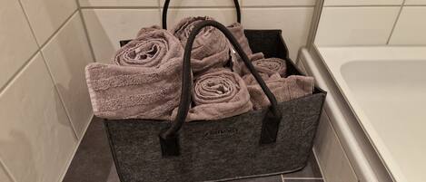 A dark gray tote bag with handles, containing several rolled towels, placed on a tiled floor in a corner.