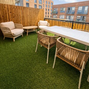 Eine Terrasse mit Kunstgrifflfläche, Korbmöbeln und Blick auf ein Ziegelgebäude.