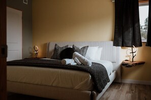 Un letto rifatto con copriletto bianco e una coperta scura, un comodino con una lampada, e una finestra con tende scure.