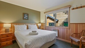 Ein Schlafzimmer mit einem großen Bett, einem Holzkopfteil, einem Nachttisch mit Lampe, einem Korbstuhl und einem Fenster mit Blick auf ein Holzhaus.