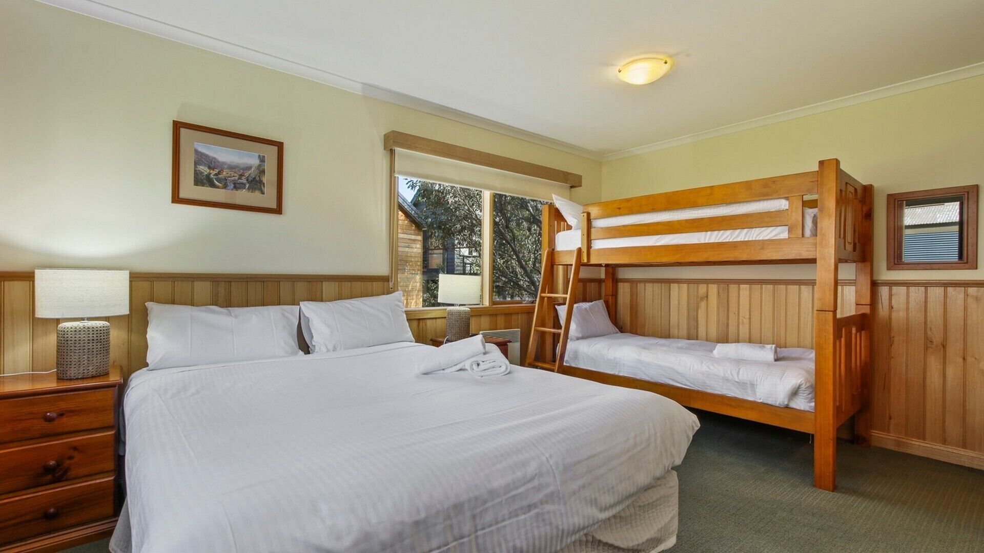 A room with a large bed, a smaller bunk bed, wooden paneling, a window with a view, a bedside lamp, and a framed picture on the wall.