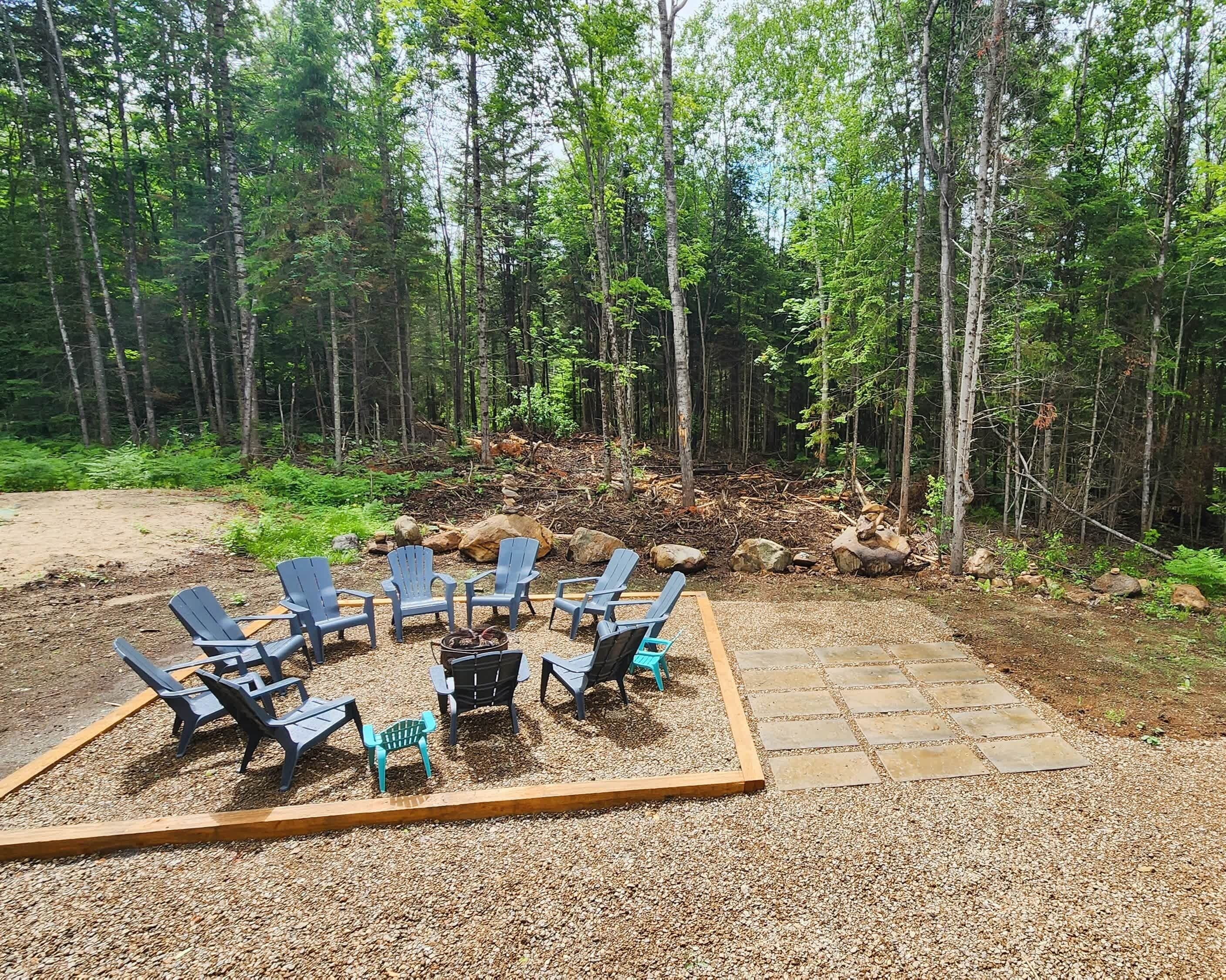 Area esterna con sedute in stile Adirondack e un braciere, circondata da un patio in ghiaia, con uno sfondo di alberi.