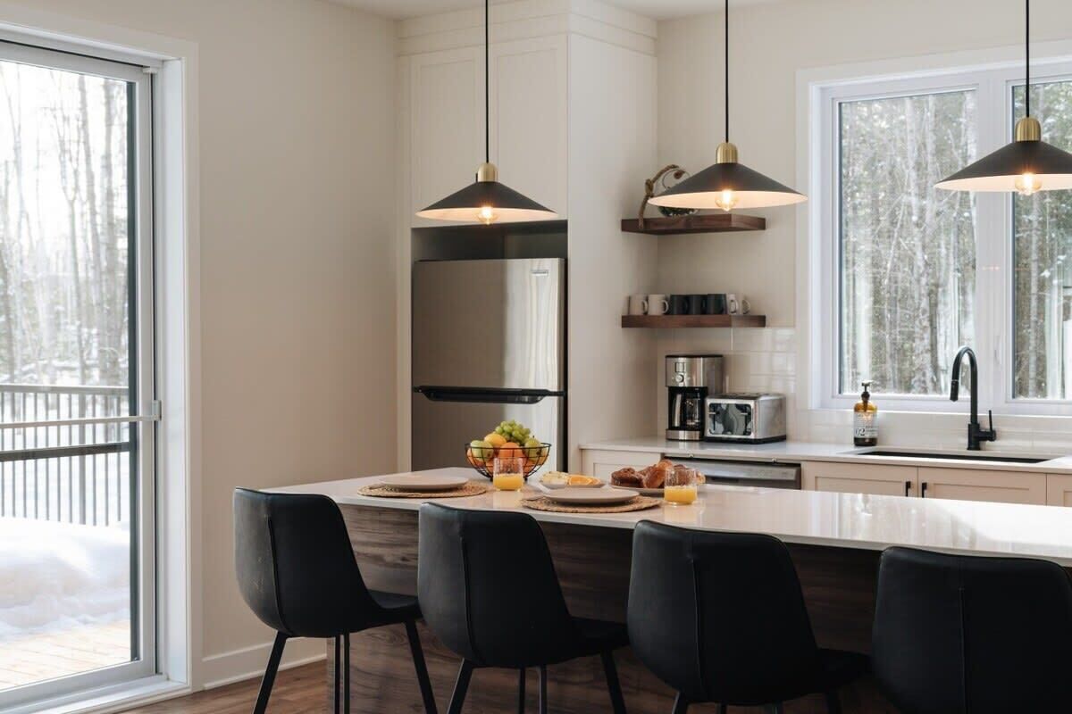 A modern kitchen with a central island, black chairs, pendant lights, a refrigerator, a coffee maker, and a window with a view of trees.