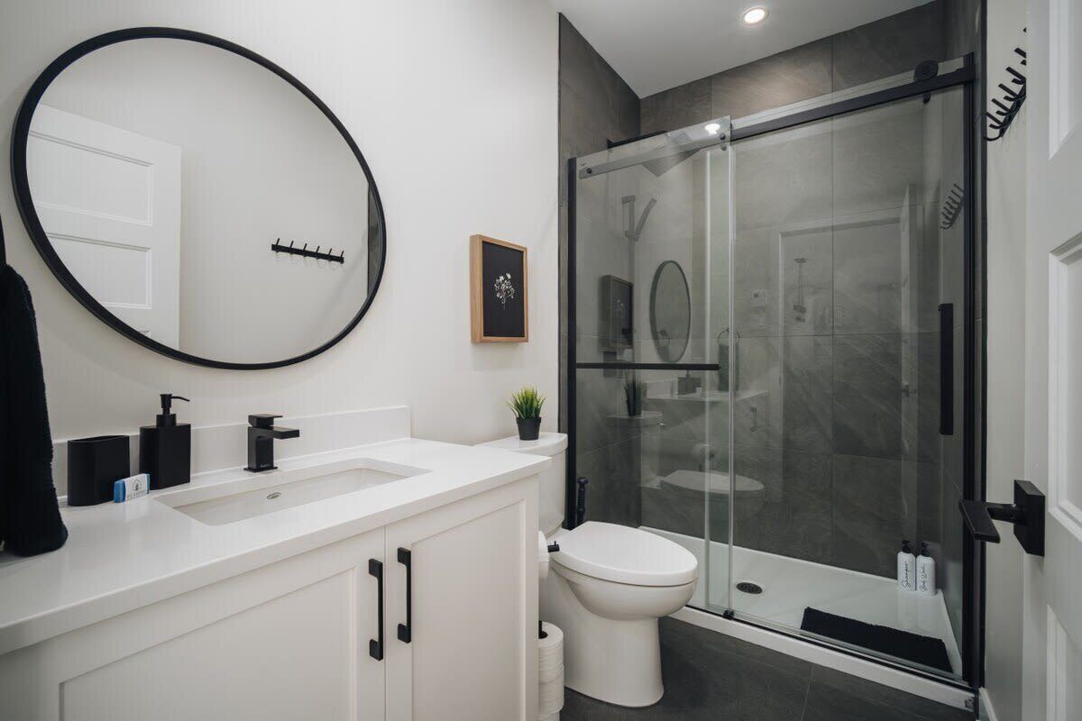 Un baño moderno con un espejo redondo grande, cabina de ducha de vidrio, lavamanos blanco y un inodoro.