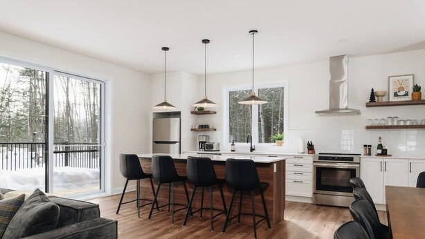 A modern kitchen with a central island, bar stools, pendant lighting, and a large window offering a view of a snowy landscape. - Peaceful Chalet Tremblant, hot tub lakes & Kayaks (Amherst)