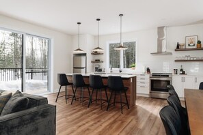A modern kitchen with a central island, bar stools, pendant lighting, and a large window offering a view of a snowy landscape. - Peaceful Chalet Tremblant, hot tub lakes & Kayaks (Amherst)