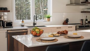 A modern kitchen with a large island, a window with a view of trees, and a sink with a faucet. - Peaceful Chalet Tremblant, hot tub lakes & Kayaks (Amherst)