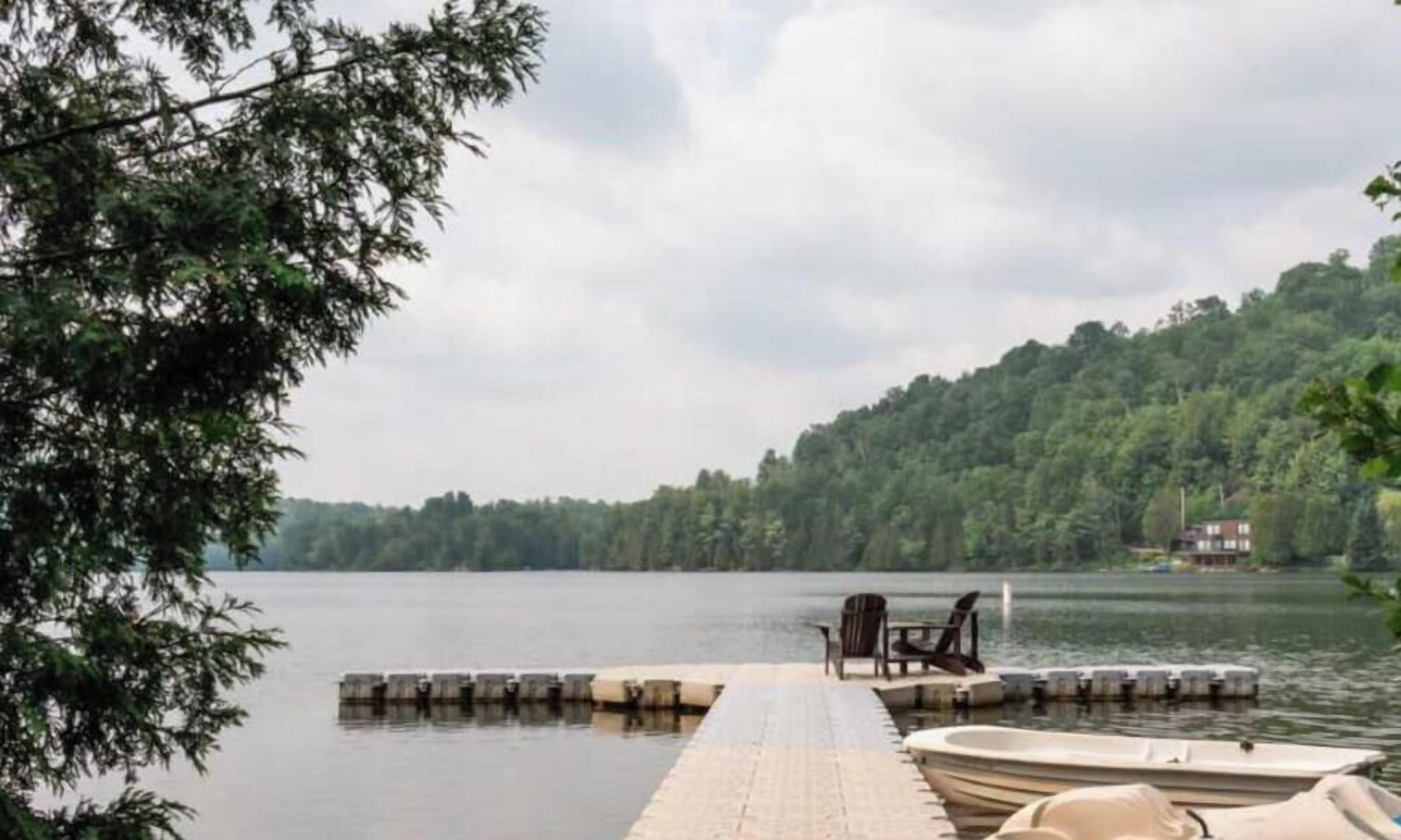 A dock with a rowboat and a chair by the water, surrounded by trees and a house in the background.