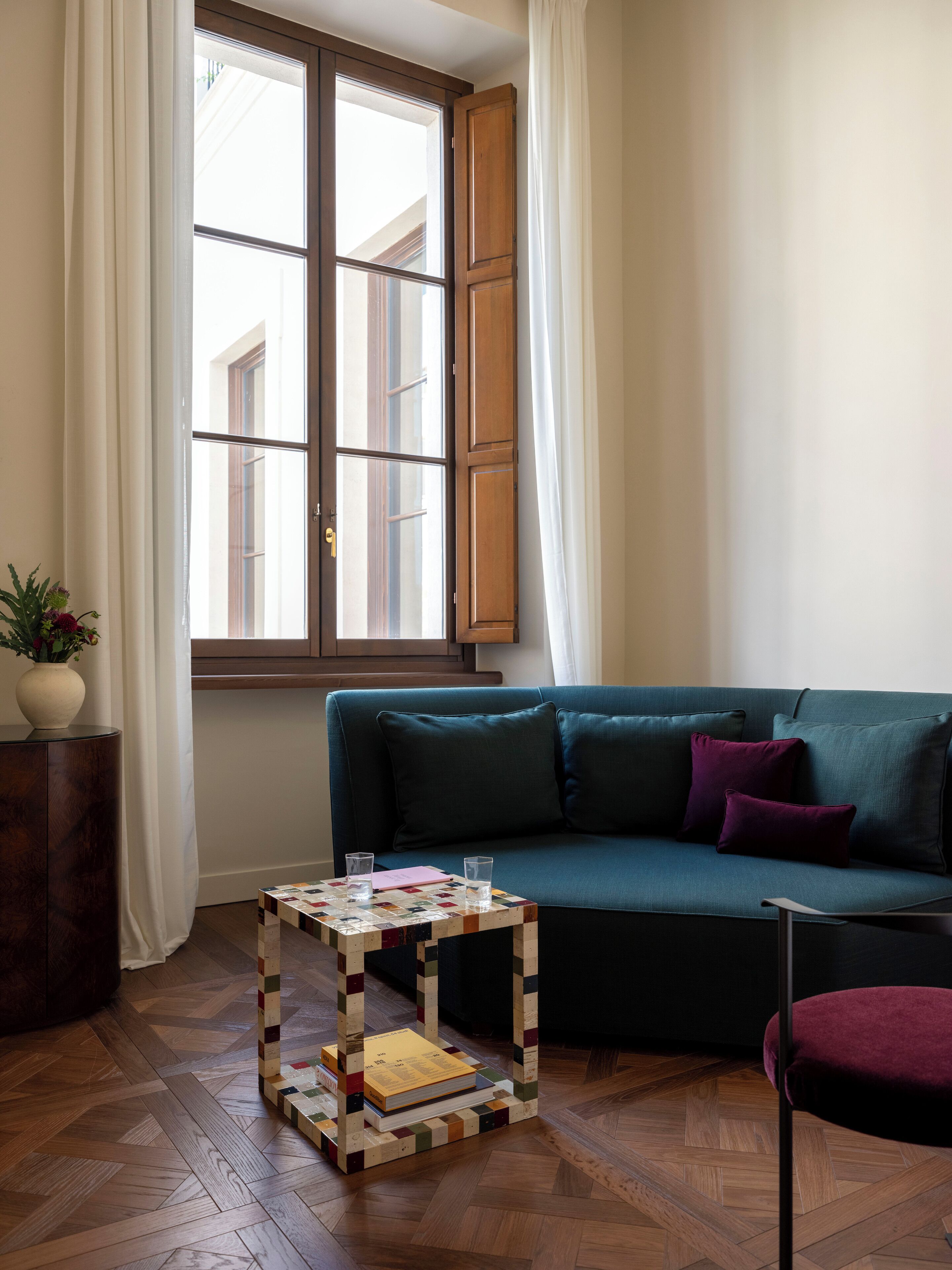 Un salón con suelo de madera en patrón de espiga, un sofá verde con cojines morados, una pequeña mesa de centro de madera con libros, y una ventana con cortinas blancas.