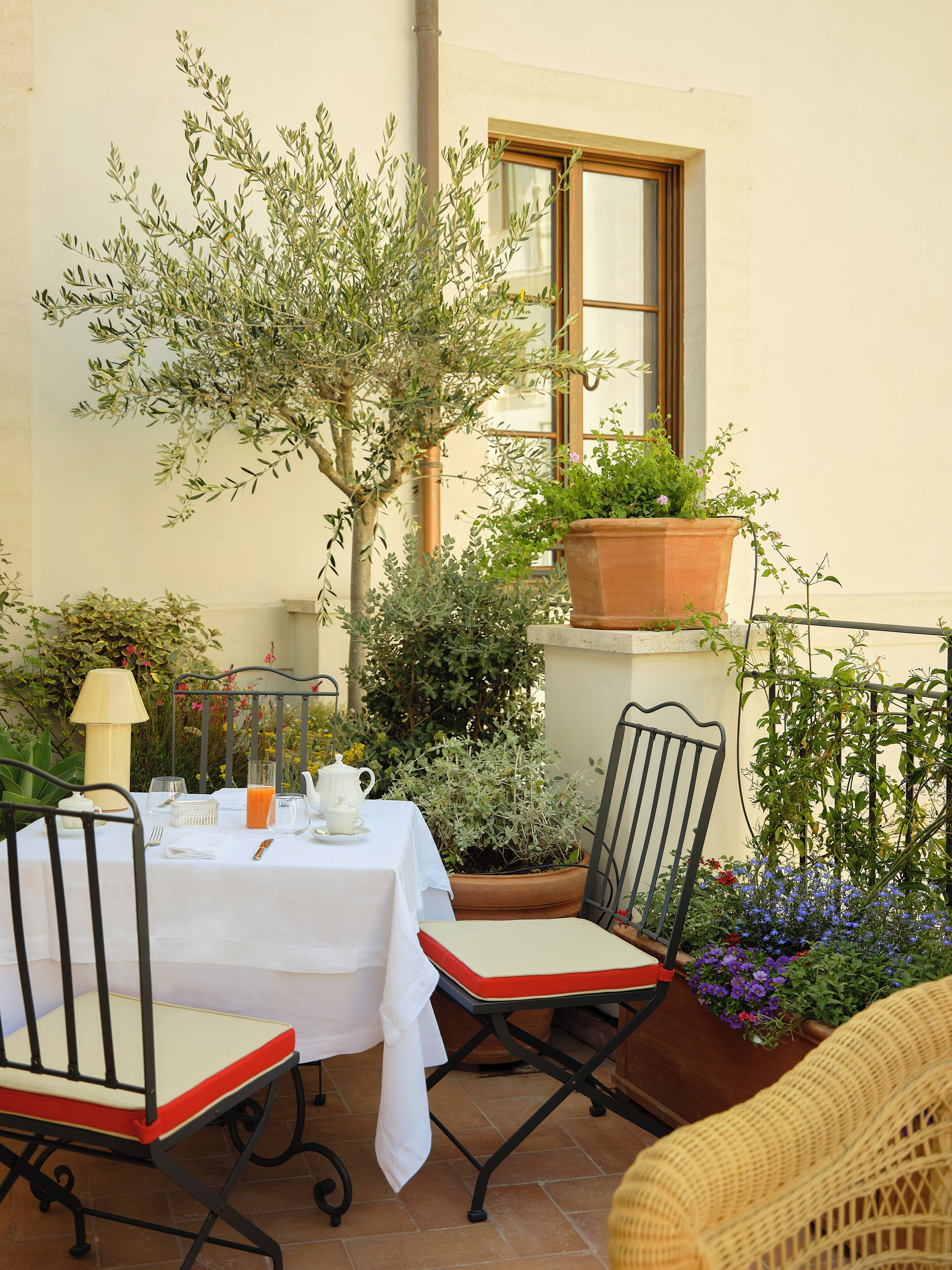 Zona pranzo all'aperto con tavolo apparecchiato per due, circondata da piante in vaso e un piccolo albero.