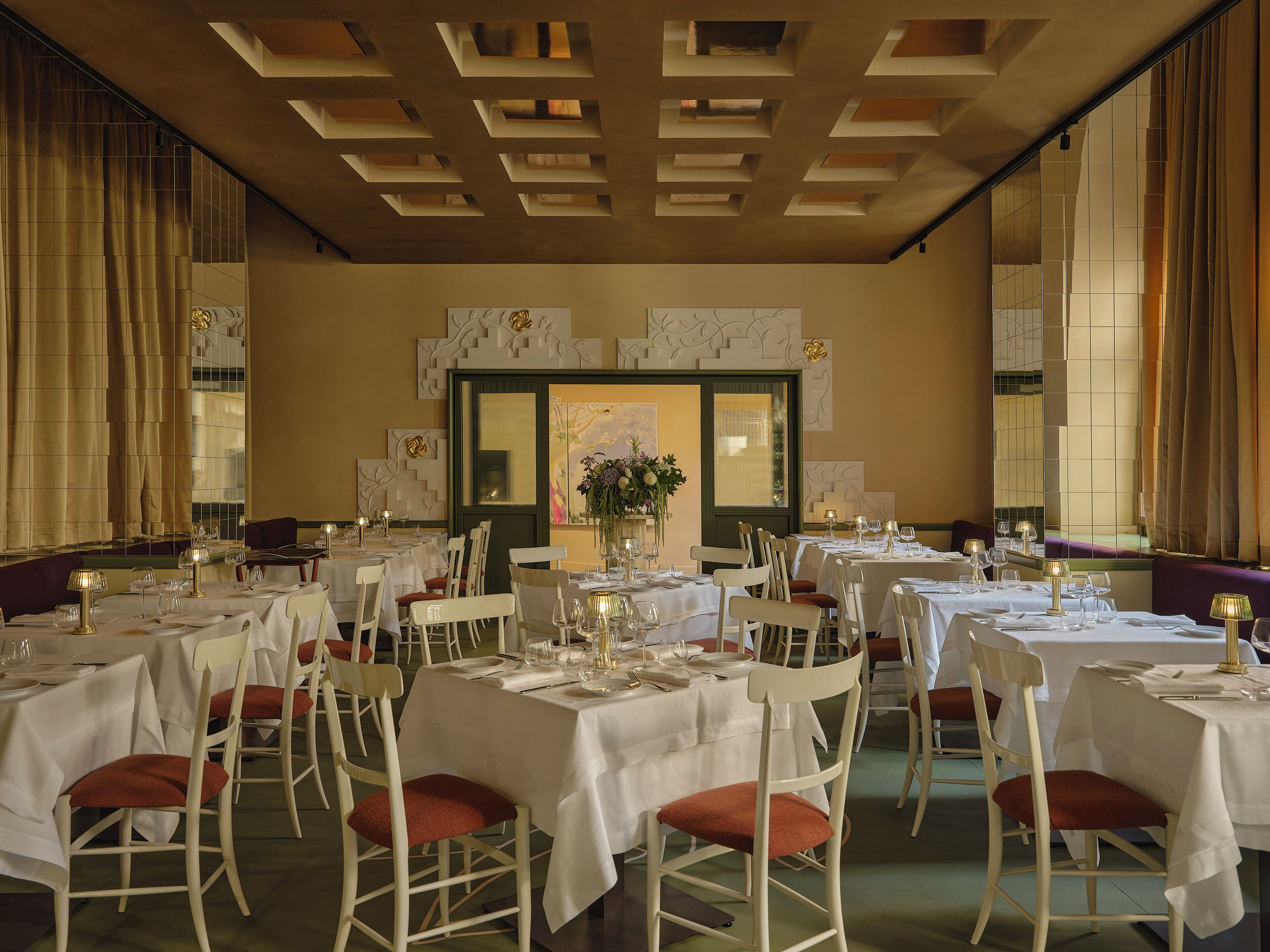 A formal dining room with multiple tables set for guests, featuring white tablecloths and red cushioned chairs.