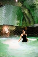 Una persona en traje de baño negro en una piscina de color verdoso, rodeada por una cerca de malla.