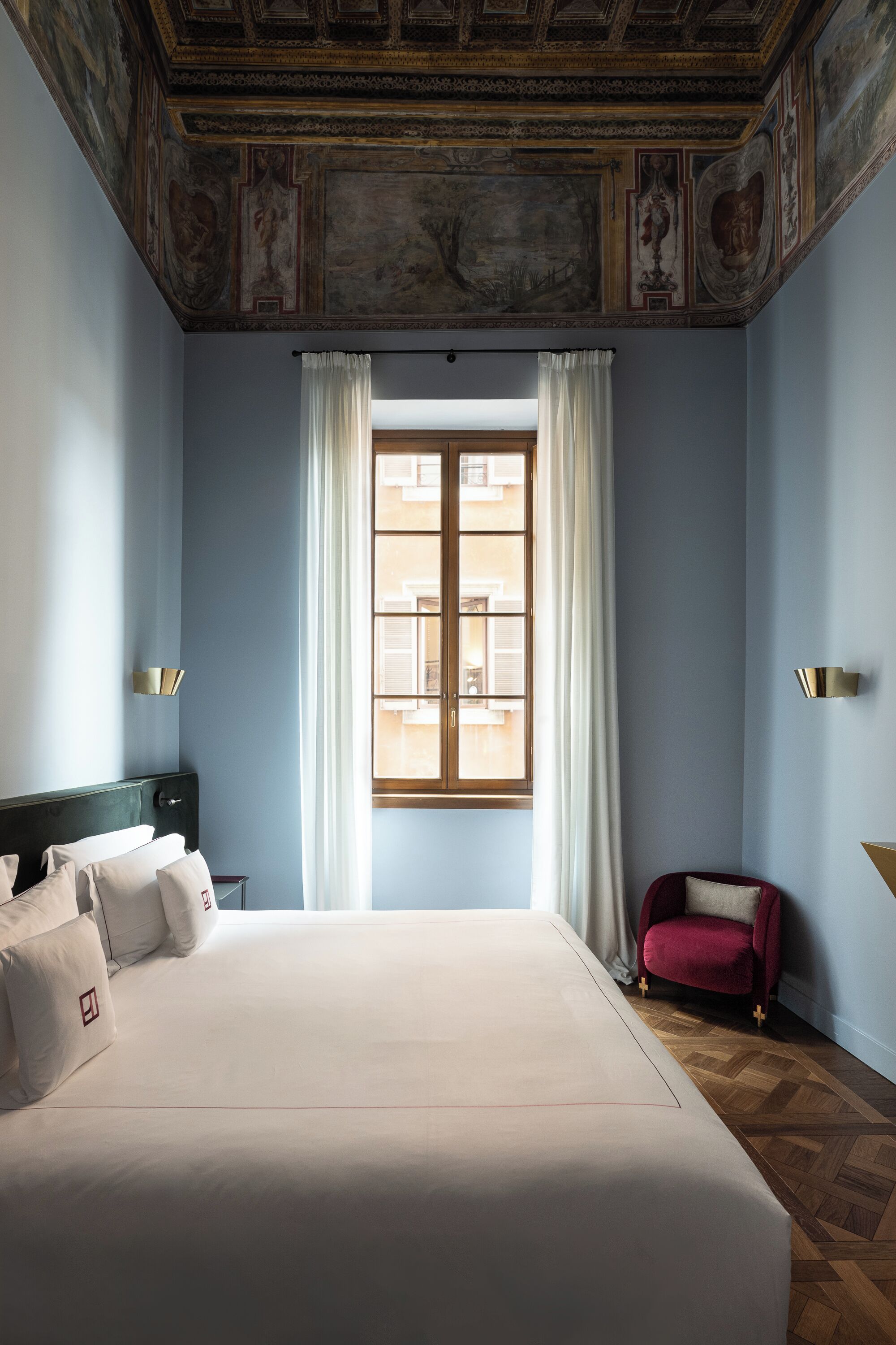 A hotel room with a large bed, a red armchair, a window with curtains, and a decorative ceiling.