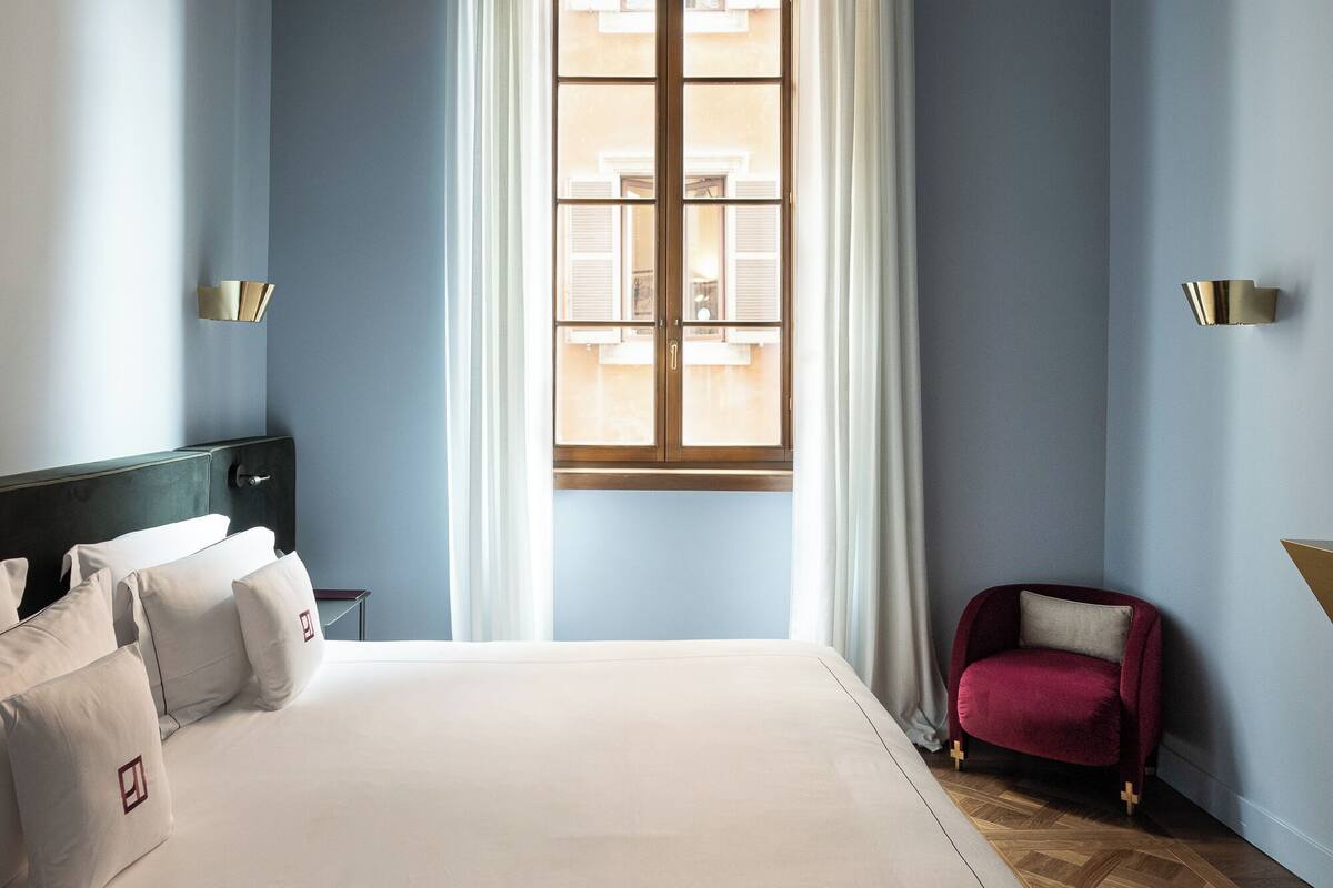 A hotel room with a large bed, a red armchair, a window with curtains, and a decorative ceiling.
