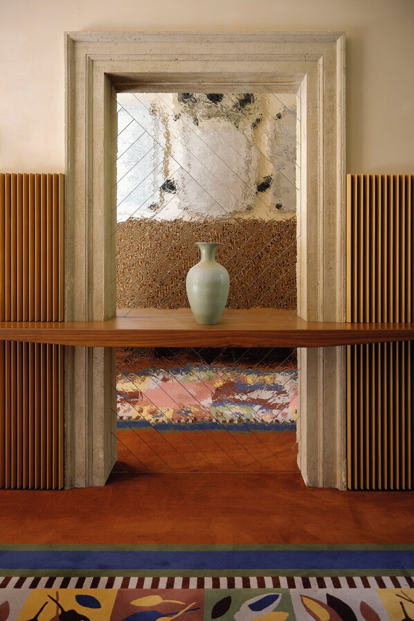 A framed mirror with a vase on a wooden shelf, a patterned rug, and a textured wall. - Palazzo Talìa - Small Luxury Hotels of the World (Rome)