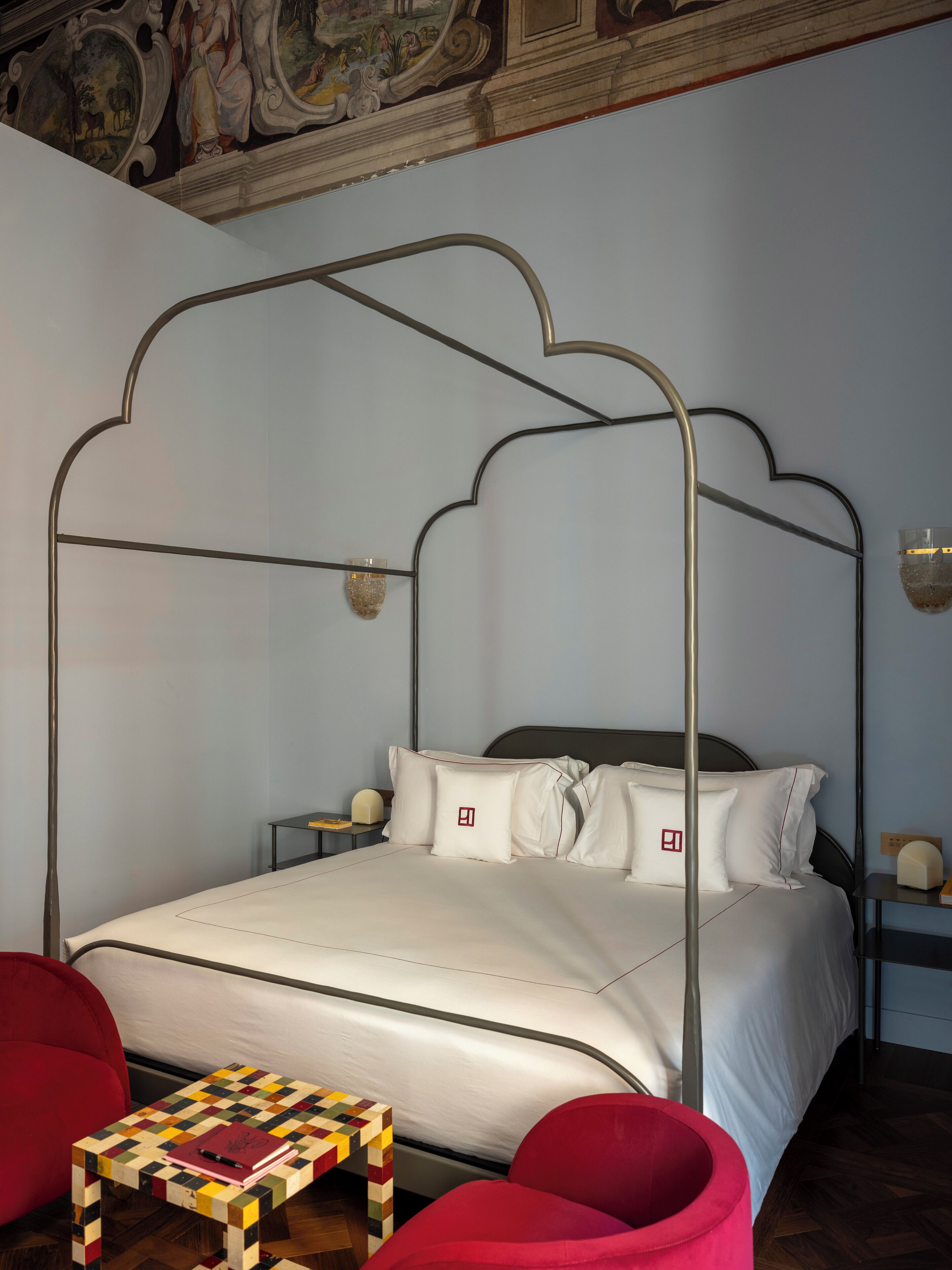 A four-poster bed with white linens, a small wooden table with a checkered top, and a red chair.