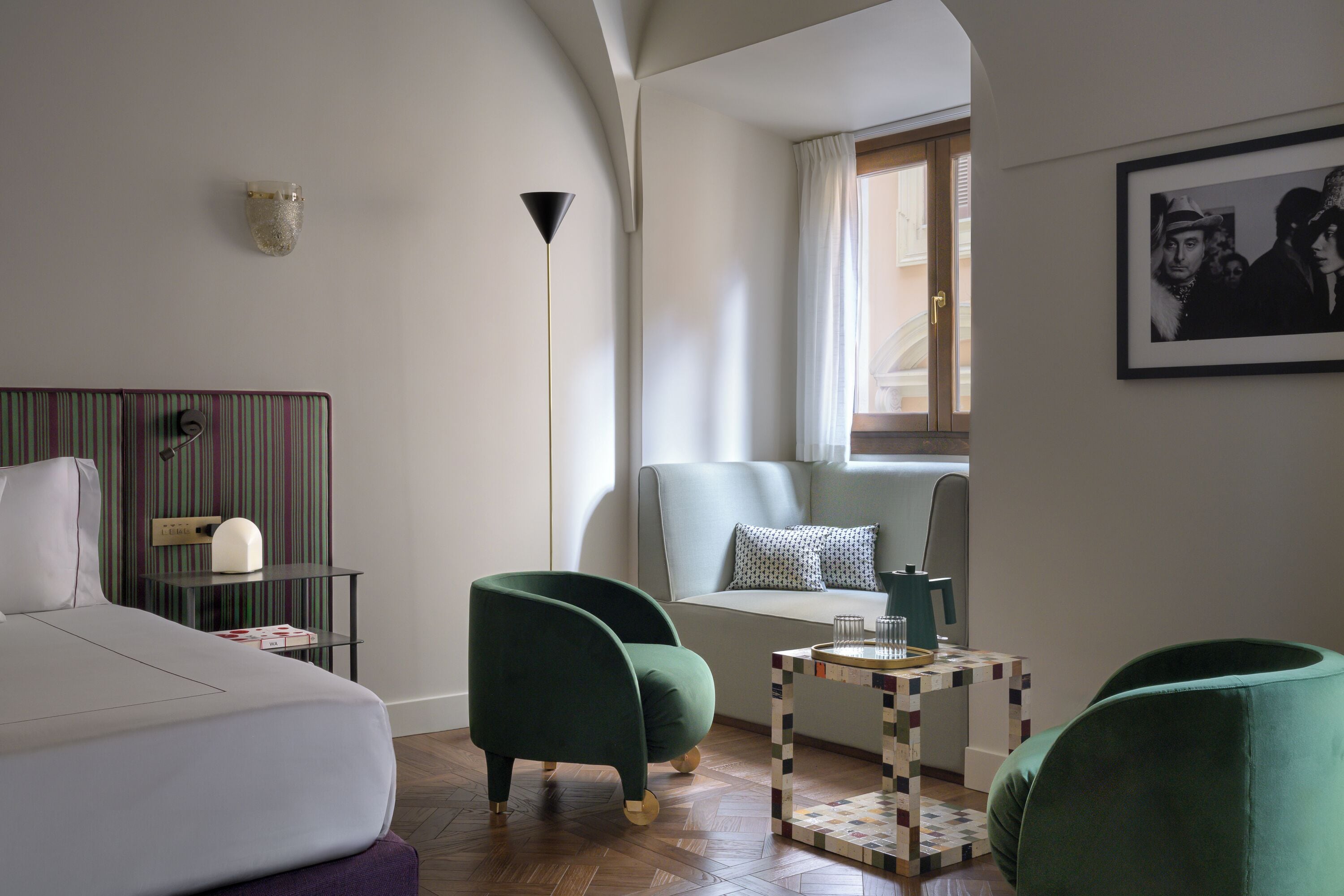 Ein modernes Hotelzimmer mit einem Bett, zwei grünen Sessel, einem kleinen Tisch mit Kaffeemaschine, einer Wandleuchte und einem gerahmten Bild.