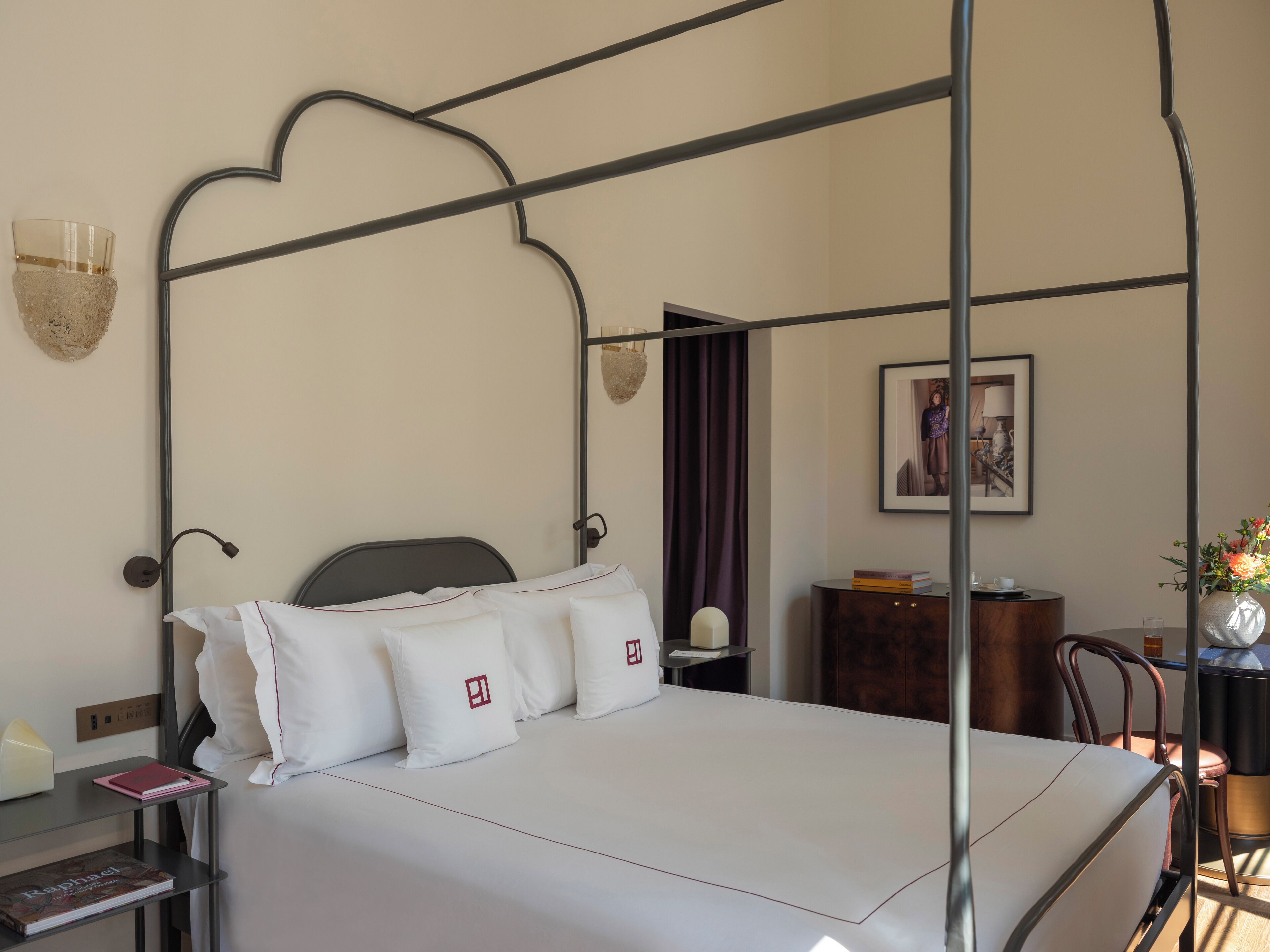 Una cama bien hecha con sábanas y almohadas blancas, una mesita de noche con un libro y una lámpara, un cuadro en la pared y una pequeña mesita de madera con un florero.