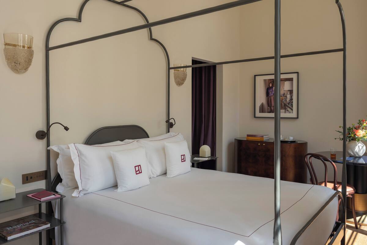 A neatly made bed with white linens and pillows, a bedside table with a book and a lamp, a framed picture on the wall, and a small wooden side table with a vase of flowers.