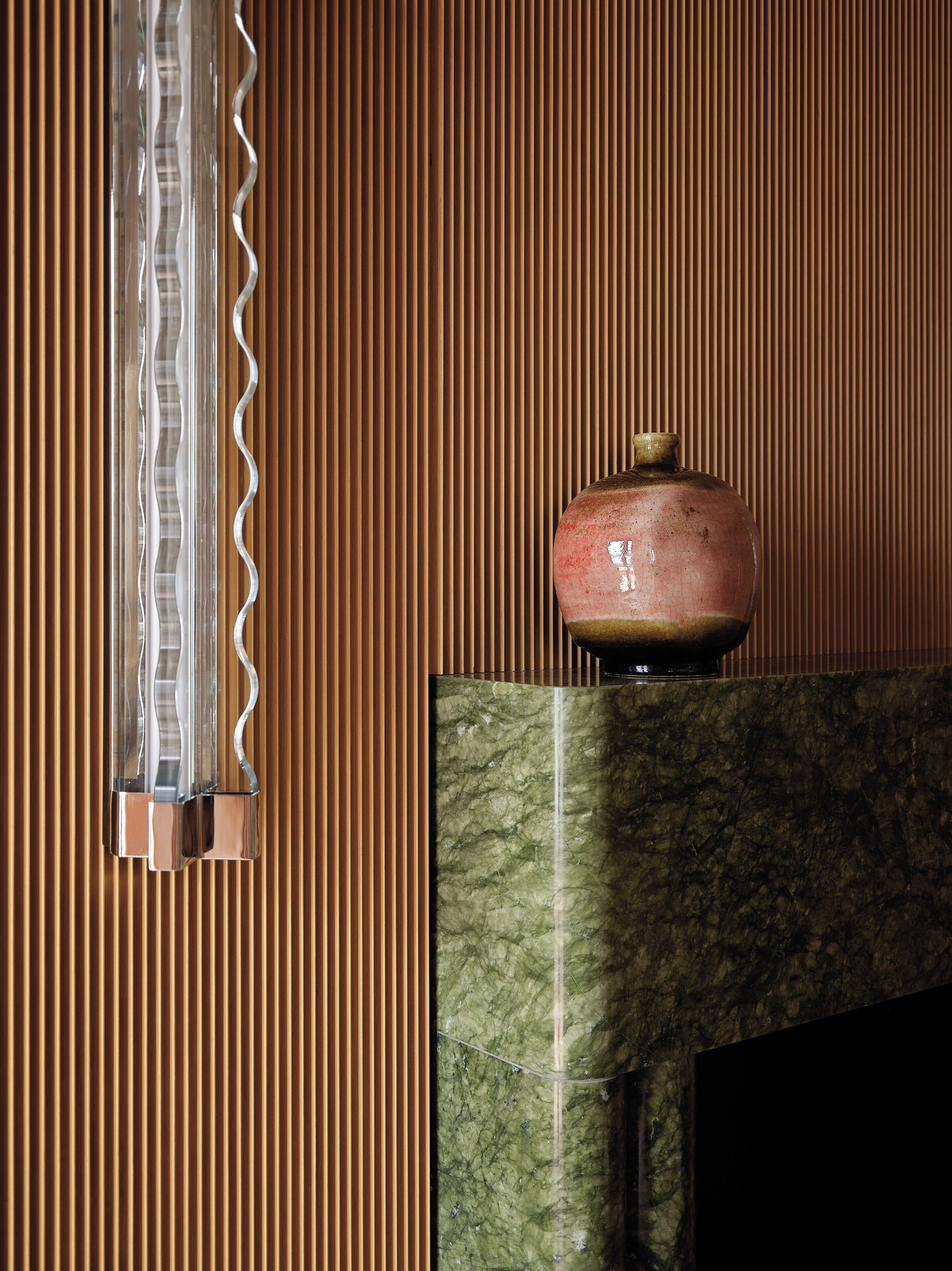 A room with a wooden slatted wall, a vase on a marble fireplace, and a clear, wavy-patterned light fixture.