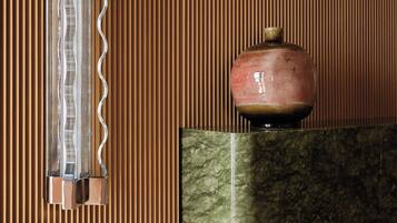 A room with a wooden slatted wall, a vase on a marble fireplace, and a clear, wavy-patterned light fixture.