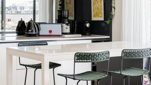 A modern kitchen with a white countertop, black cabinets, and a coffee machine. There are three green chairs around the table, and a plant is on the wall. - StayCentral - The Stanley@Collingwood - Smith St, Collingwood - 2 Bedrooms, 2 Beds, 1 Bathroom1, 1 secure undercover parking spot. (Collingwood)