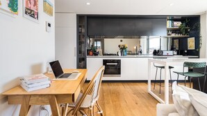 A modern kitchen with a central island, white cabinetry, and a dining area with a wooden table and chairs. - StayCentral - The Stanley@Collingwood - Smith St, Collingwood - 2 Bedrooms, 2 Beds, 1 Bathroom1, 1 secure undercover parking spot. (Collingwood)