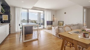 A modern apartment with a kitchen, dining area, and living room. It features a large window, a flat-screen TV, and a laptop on the dining table. - StayCentral - The Stanley@Collingwood - Smith St, Collingwood - 2 Bedrooms, 2 Beds, 1 Bathroom1, 1 secure undercover parking spot. (Collingwood)