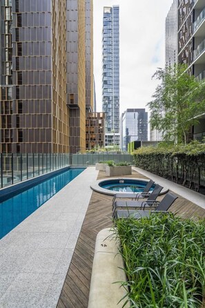 Un espace piscine aménagé sur le toit, avec une piscine rectangulaire et un jacuzzi, le tout entouré d’immeubles modernes et de verdure.