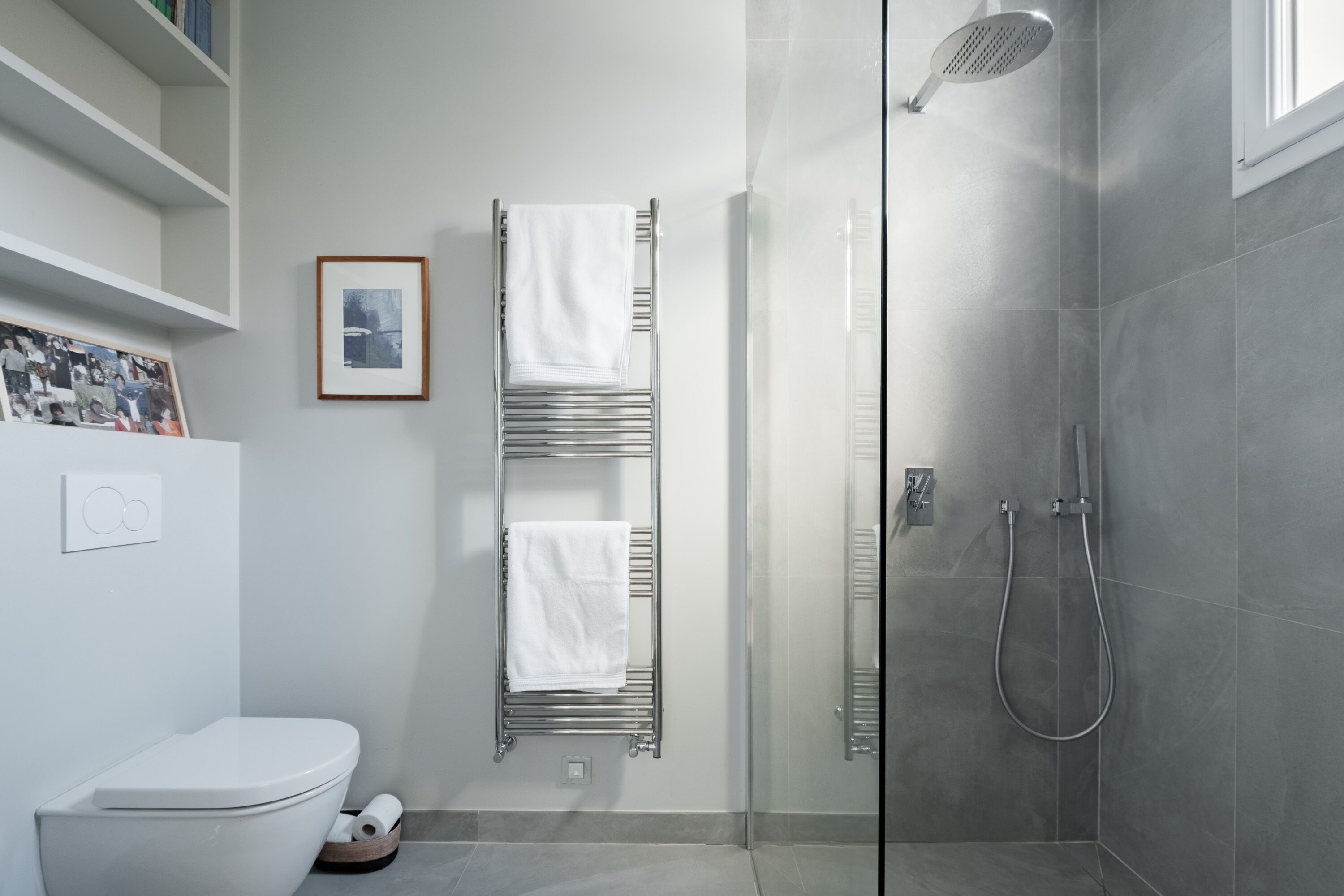A modern bathroom with a toilet, a towel rack with three towels, a shower with a glass door, and a framed picture on the wall.