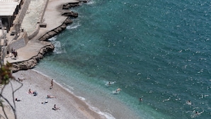 A coastal beach with clear turquoise water, rocky shoreline, and people sunbathing and swimming.