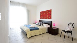 A bedroom with a bed, two bedside tables, a chair, a window with curtains, a radiator, and a wall with a colorful painting. - Trilo Mina 3 in Gabicce Mare (Cattolica)