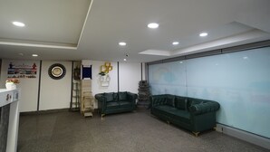 Un área de espera moderna con dos sofás de cuero verde, una pared blanca con un reloj y un cartel enmarcado, una planta pequeña y un mamparo de vidrio.