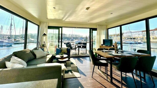 A spacious living area with a large window overlooking a marina, a dining area with a table and chairs, and a sofa with cushions.
