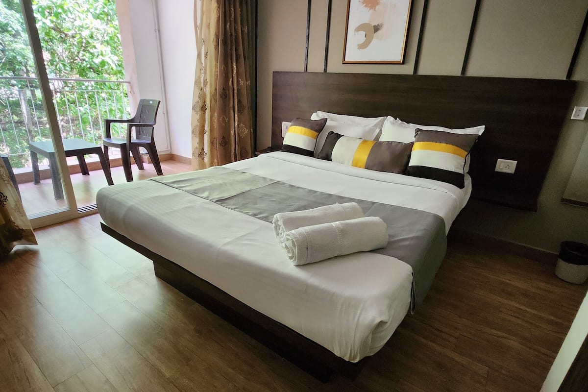 A modern bedroom with a large bed, a balcony with a chair, and a view of greenery.