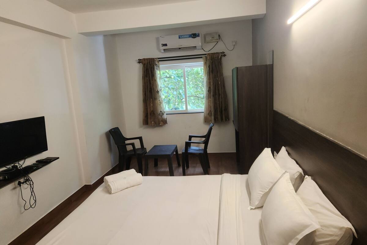A hotel room with a bed, a TV, a desk, two chairs, a window with curtains, and an air conditioning unit.