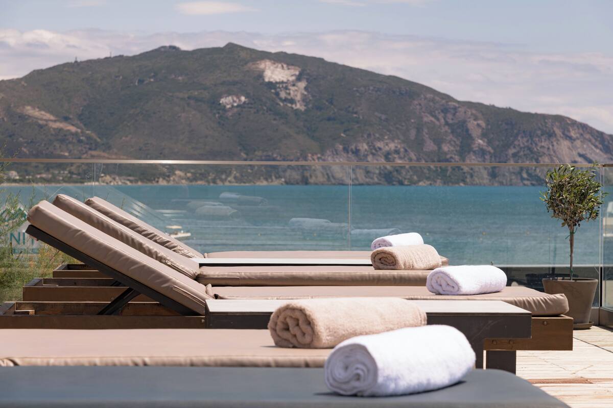 A terrace with sun loungers and towels, overlooking a body of water and a mountainous landscape.